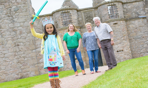 A young girl in a blue dress and stripy rainbow socks has a crown on her head and holds a plastic sword above her head. Behind her and in front of an old castle, are mum and grandparents. Everyone is smiling. 
