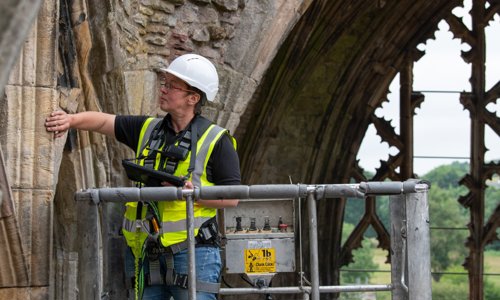 A person standing in a cherrypicker wearing protective workwear inspects a stone arch in a medieval abbey.