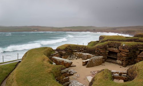 Breaking waves on a stormy day hit a beach just behind a series of archaeological excavations in the foreground.