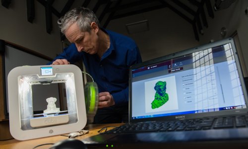 A person uses a 3D printer to create a small white model of a historic figure. The figure can also be seen on a laptop computer in the foreground.