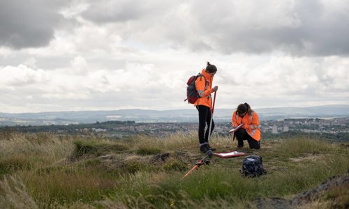 Two people in outdoor gear working with survey equipment on a windy looking moor. One is standing, the other kneeling down and writing something on a clipboard.