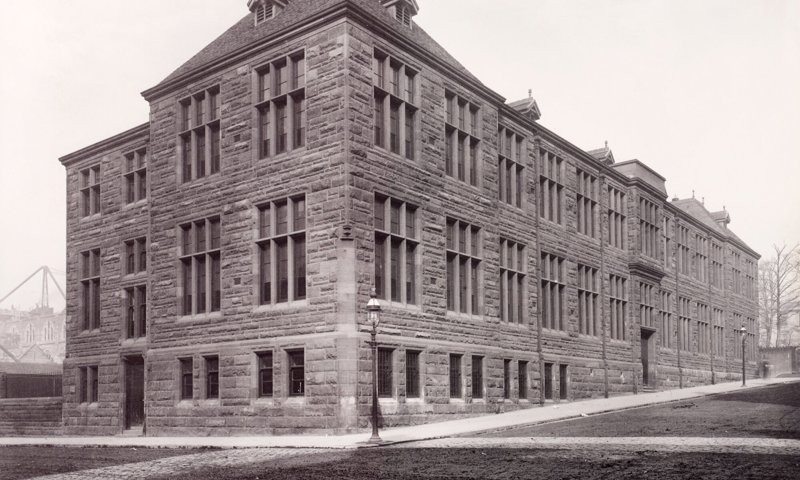 A black and white photo of a three-story angular school building, made of brick and stone and with large vertical windows on both sides. The school building is surrounded by bare earth and clean footpaths, as if it has been recently finished.