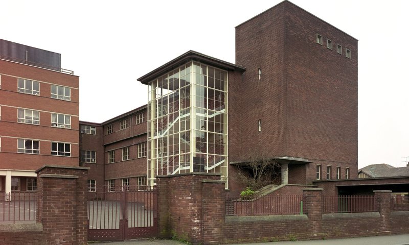 A large, angular brick and glass secondary school, with tall brick sides and an open school yard. The building is separated from the road by a stone wall and gate.