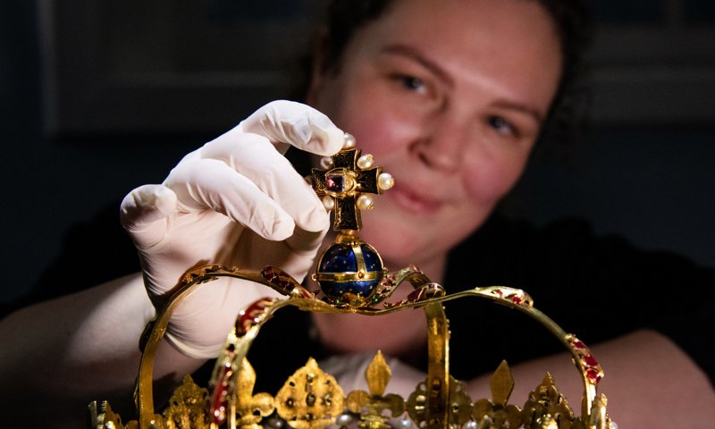 A person wearing a latex glove on their right hand touching the uppermost part of an ornate crown, made of gold and studded with many pearls and precious jewels.