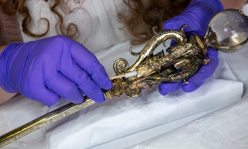 A person wearing blue gloves using a cotton bud to clean a jewelled sceptre, which is made of silver and has a large crystal sphere at its top.