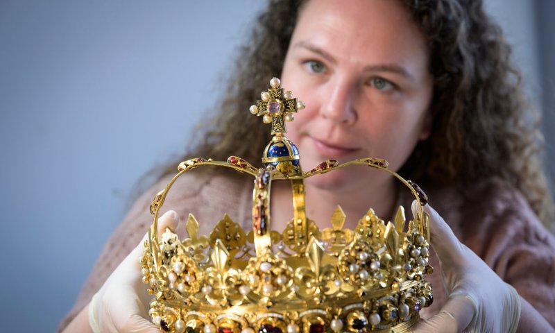 A person wearing gloves looks closely at a golden crown, set with many jewels and precious stones.
