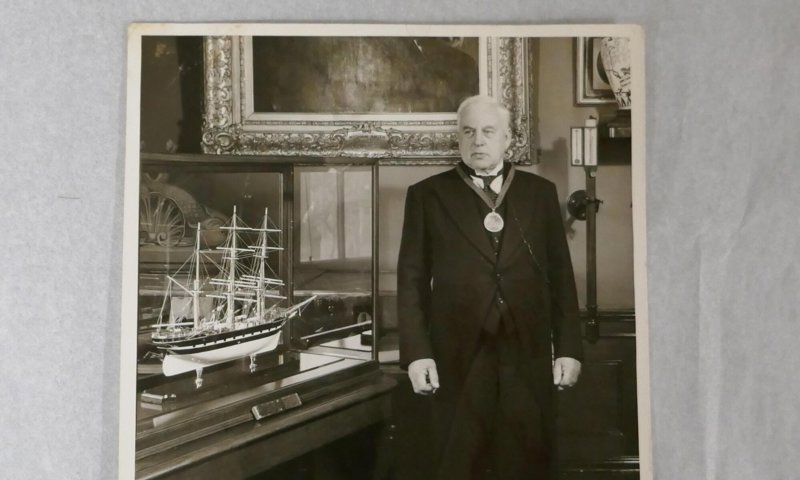 A person wearing a dark suit and trousers stands next to a large model of a wooden sailing ship in a museum. The model is behind glass, and sits on a table in front of large paintings on the wall. The person has their hands by their sides and over their suit, they are wearing a gold medal around  their neck.