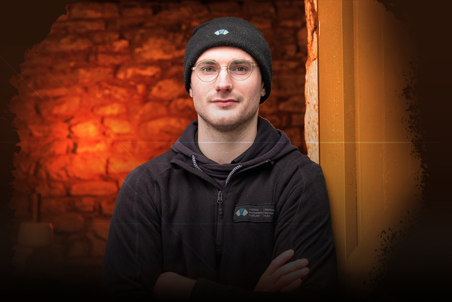 A man wearing an HES fleece and hat, leaning against a brick wall 