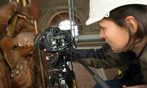 A person wearing a hard hat looks into a digital SLR camera which is pointing at a metal statue of a person in armour.