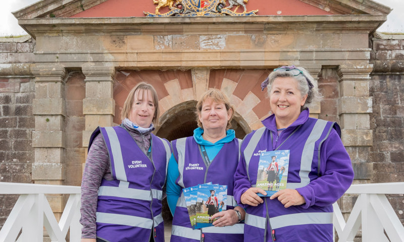 Three people standing on a drawbridge wearing purple high vis jackets with 'Event volunteer' written on them 