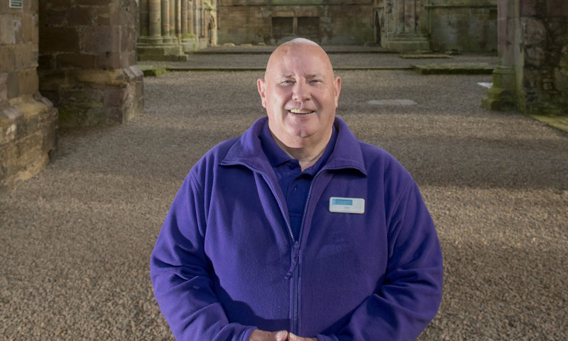 A volunteer wearing a purple HES fleece standing in the grounds of Melrose Abbey