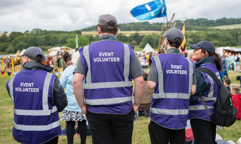 Four volunteers wearing purple high vis 'event volunteer' jackets looking on at an event taking place 