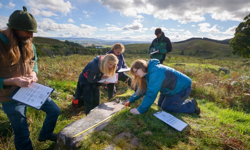 Image shows five people recording details of a metre long stone in the countryside. Some of the people are measuring the stone with a tape measure while others write down details. They are all wearing outdoor gear, and the sky is sunny with white clouds.