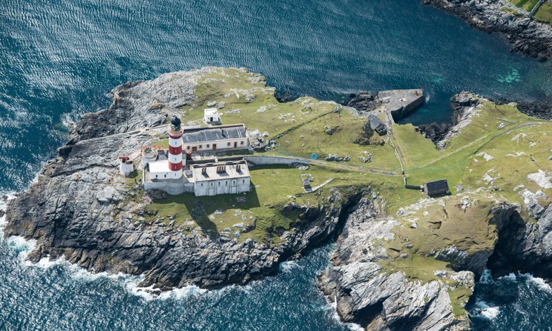 Image is an aerial photograph of a lighthouse on a rocky headland. The lighthouse is red and white striped and surrounded by smaller white buildings. The island is covered in green grass and is surrounded by the dark grey coloured sea.