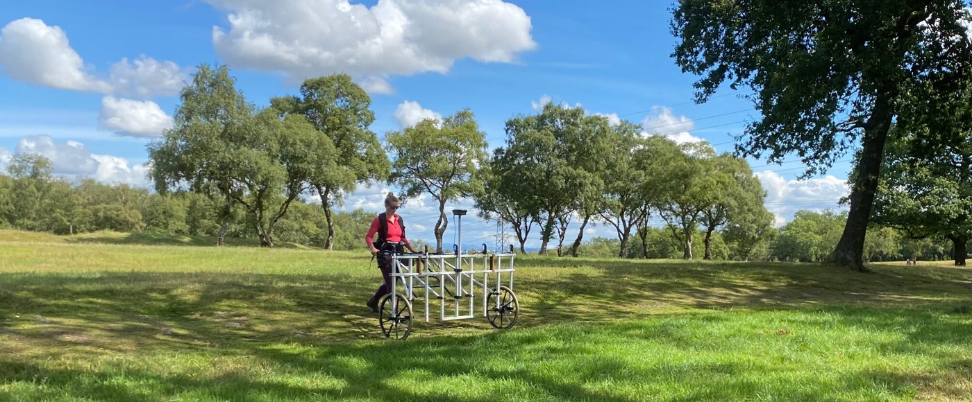 Image shows a person wearing outdoor clothing, standing behind and pushing a piece of scientific equipment across a grassy area with trees behind. The equipment is a large white lattice, about the size of a door on it side, attached to wheels.