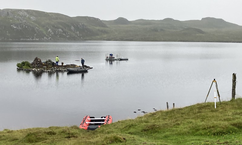 People standing on a very small island in a loch. There is scientific survey equipment on the island and the shore, with two small boats also