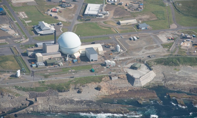 Aerial photograph of a nuclear power station and related buildings. The main building has a large round sphere next to a chimney. The complex is by the sea, with waves breaking on a rocky coastline.