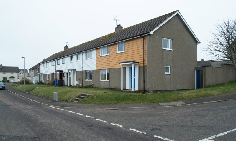 A row of semi-detached housing in Scotland. The houses have grey harling and wooden frontages, with a grey gable end and tiled rooftop. The street has grass between the houses and the pavement.
