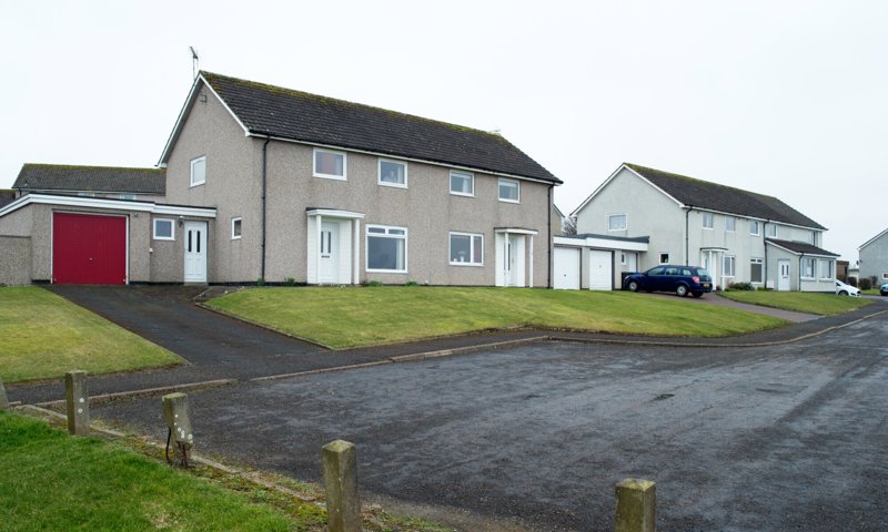 A group of semi-detached houses on an estate in Scotland. The houses are grey in colour with attached garages, and are set back from the road. In the foreground is a turning circle for vehicles to safely turn at the end of the street.