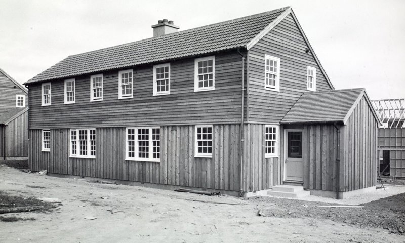 Black and white photo of a timbered house with a tiled roof. The house is in the final stages of completion and is surrounded by others with their frames showing. It has many windows with six across the first floor.