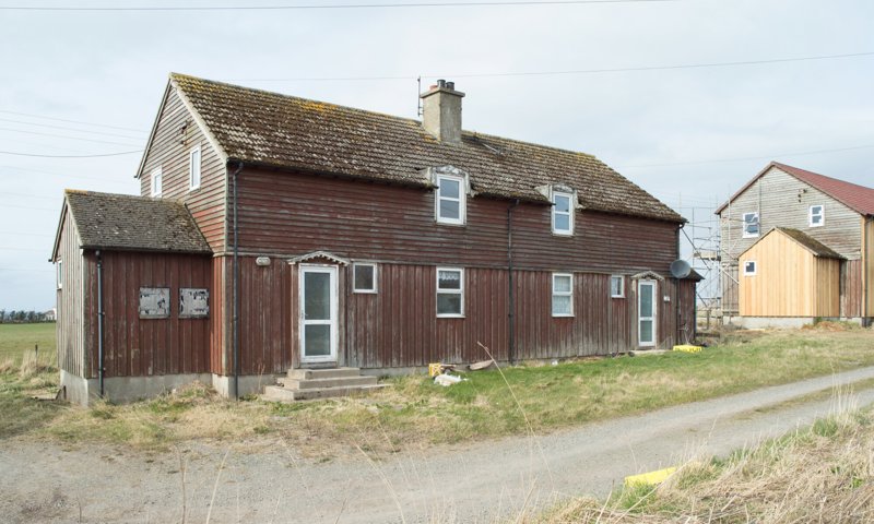 A semi-detached timber farm cottage with a slate roof. The building overlooks a gravel road and there is another double cottage next door.