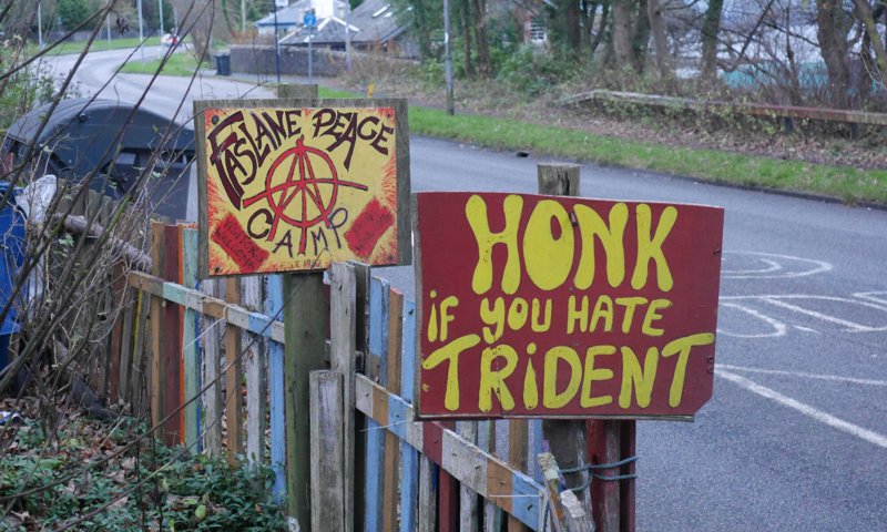 Two hand painted signs by the side of a road. The signs say Faslane Peace Camp and Honk if you hate Trident.