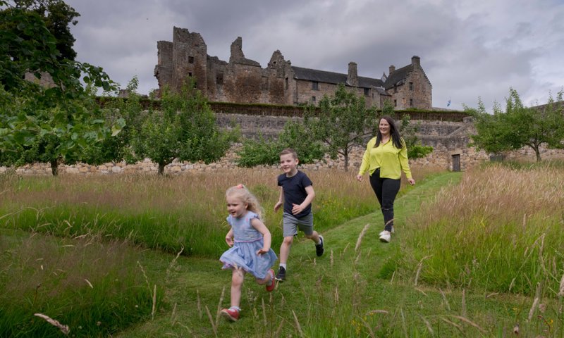 A person with two young children watches as they run ahead through a garden, with a castle in the background.