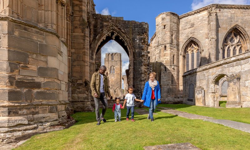 A family of two people and two children hold hands as they walk between ruined columns of an old medieval abbey.