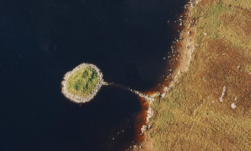 An aerial photograph taken directly above, of a small circular island surrounded by dark looking water. There is a faint line of stones visible in the water connecting the island to the rocky shore.