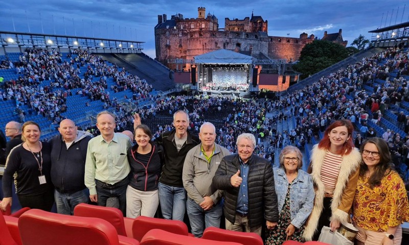 people standing in a line with the exterior of Edinburgh Castle behind them and the tattoo stands enjoying a concert